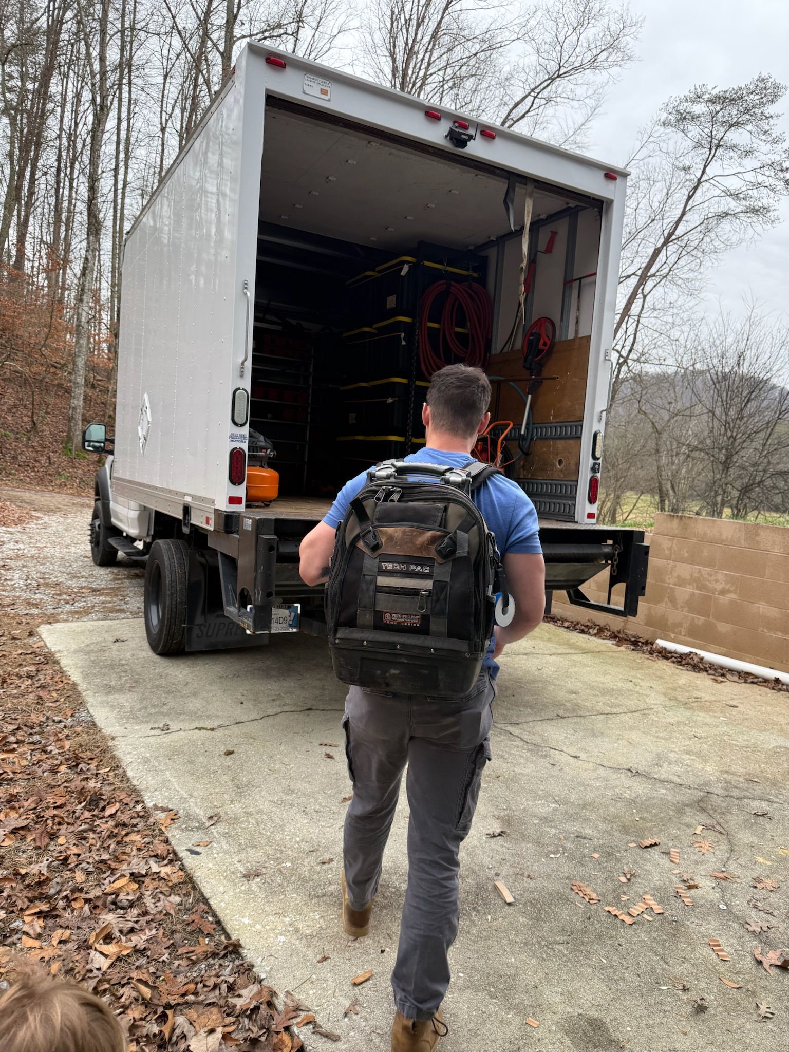 Man with backpack walks toward a large truck with open cargo doors, on a concrete driveway.