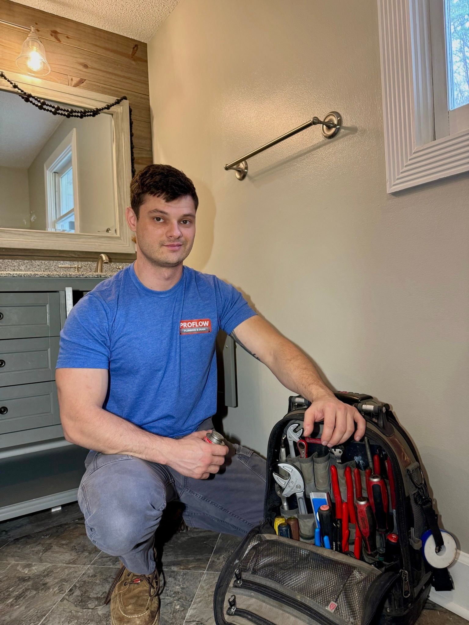 Man kneeling next to open tool bag in a bathroom, smiling. He wears a blue shirt, holding a tool.