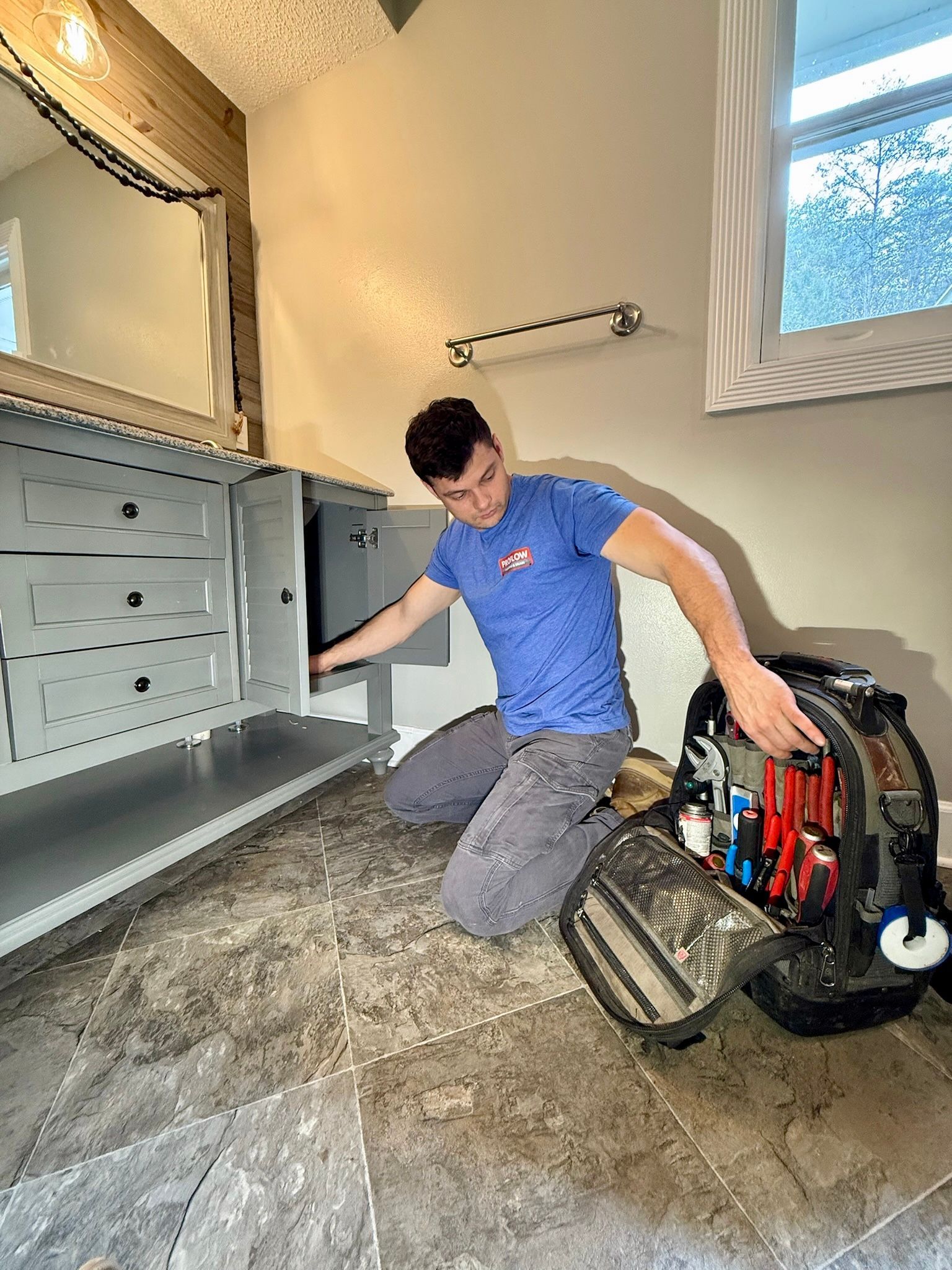 Plumber working on bathroom cabinet. He kneels near a toolbox, light blue shirt, gray pants.