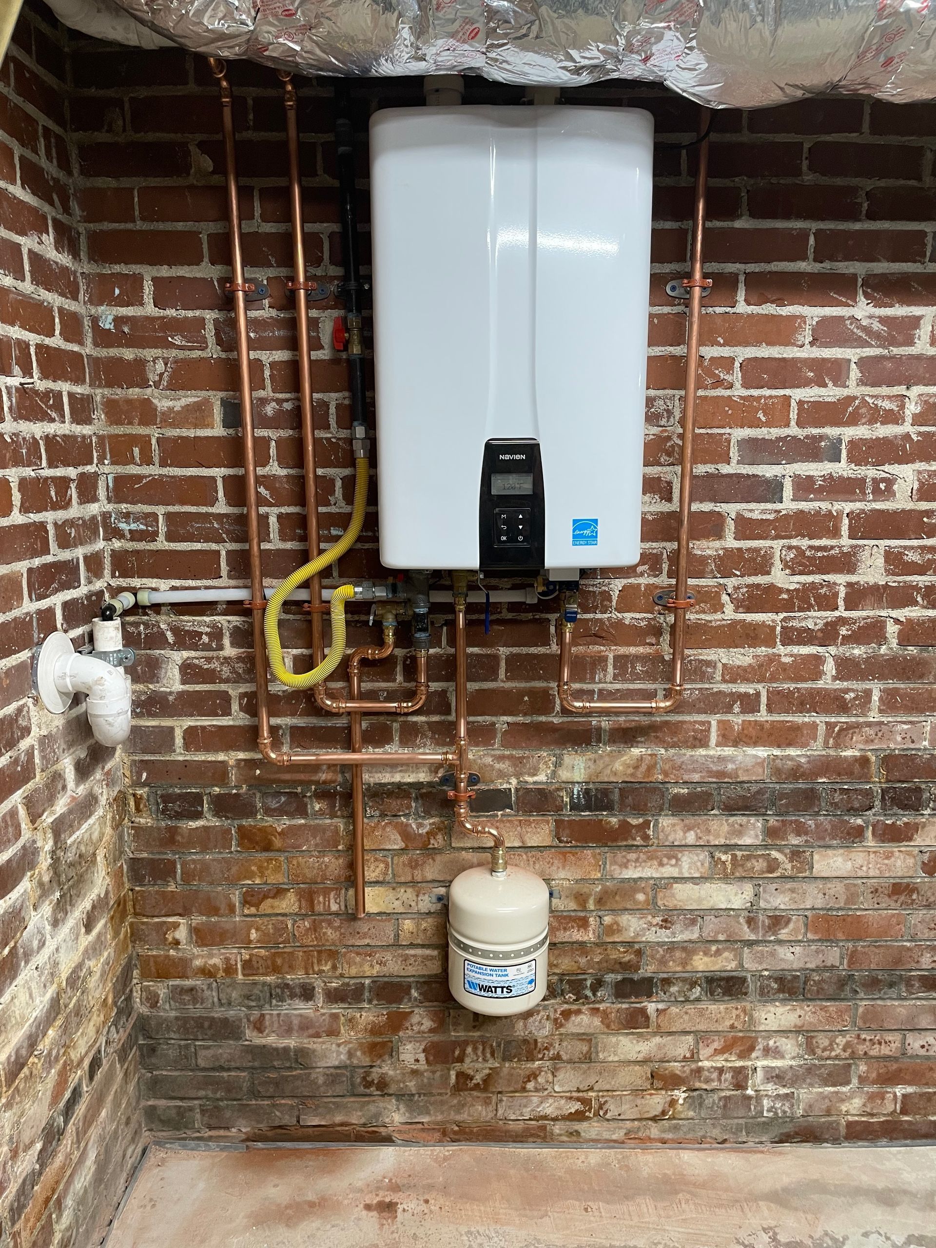 Tankless water heater installed on a brick wall with copper piping and a white expansion tank.
