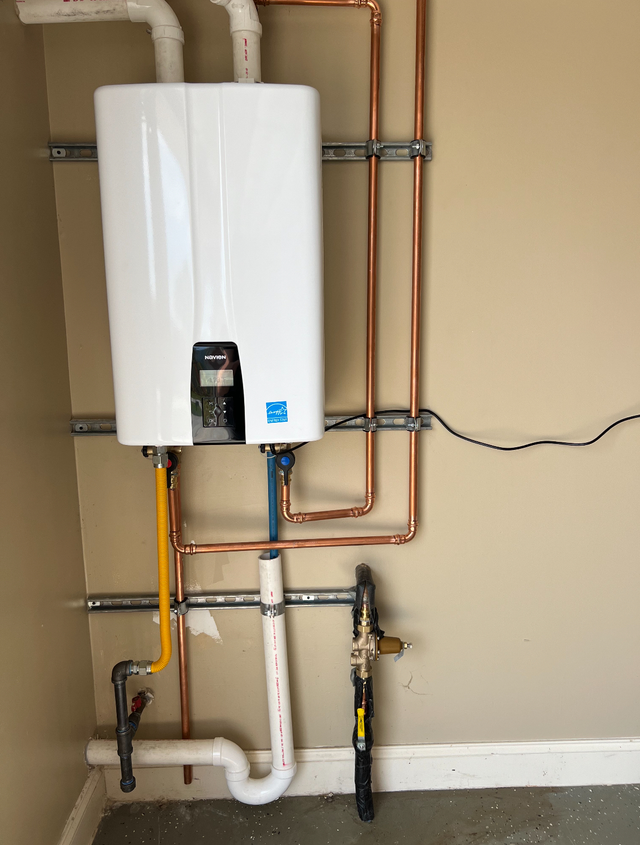 White, wall-mounted tankless water heater with copper pipes and gas line against a beige wall.