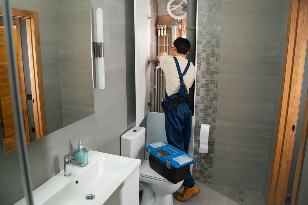 Plumber in blue overalls working on pipes in a bathroom, standing on a toilet with toolbox.