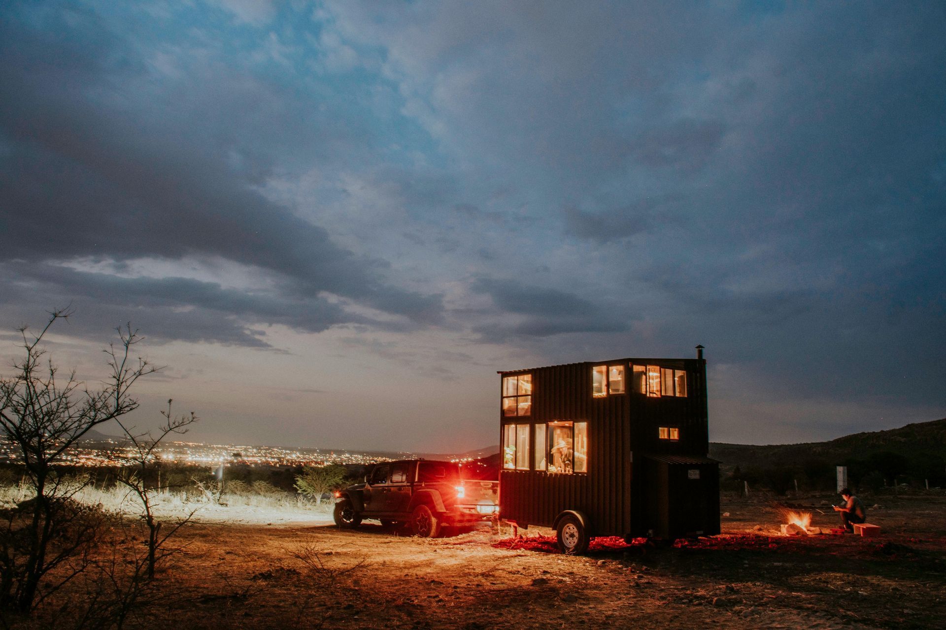 Tiny house on trailer at dusk, lit windows. Car connected, campfire nearby, city lights in distance.