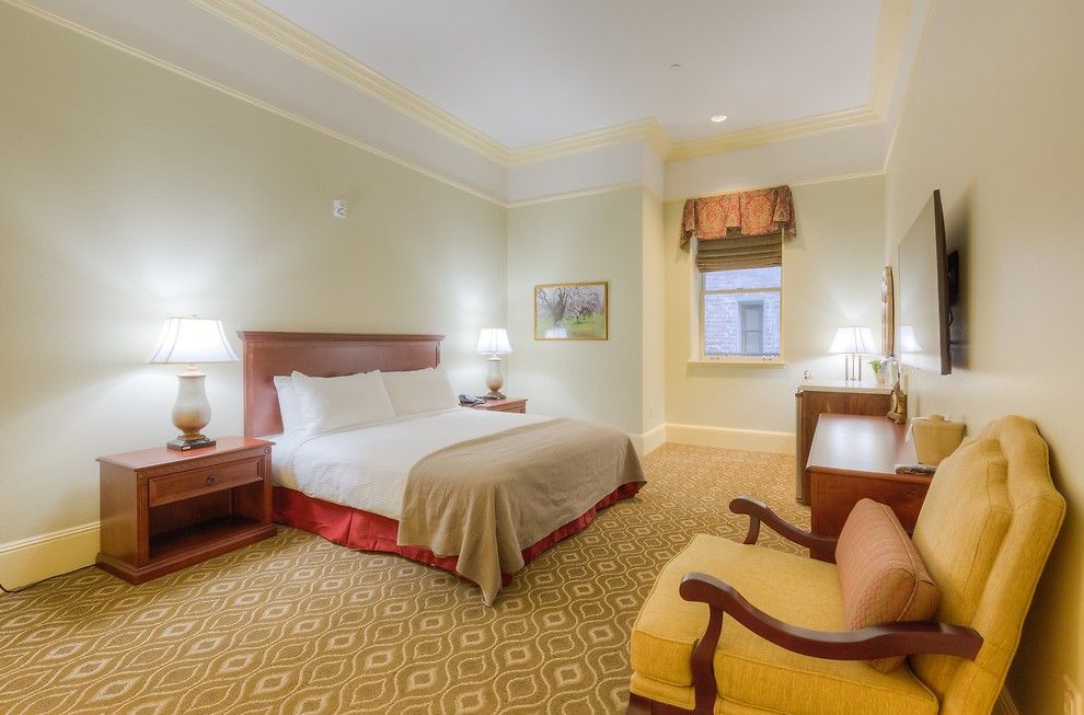 Hotel room with a queen bed, two nightstands, a yellow armchair, and a TV. The room has beige walls and patterned carpet.