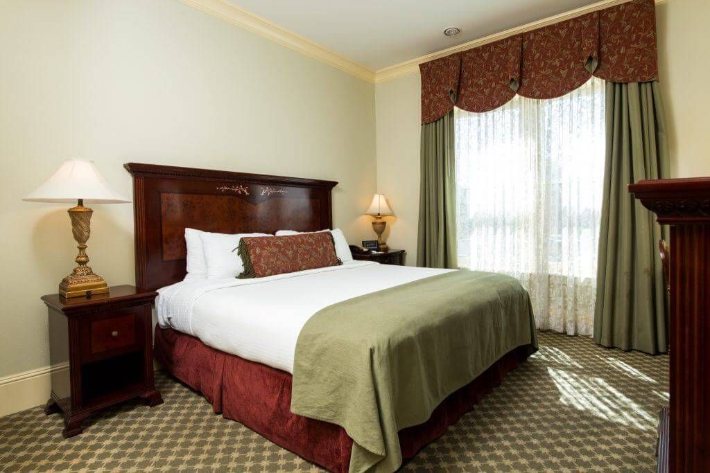 Hotel bedroom with a queen bed, nightstands with lamps, and a window with curtains. The decor is traditional.