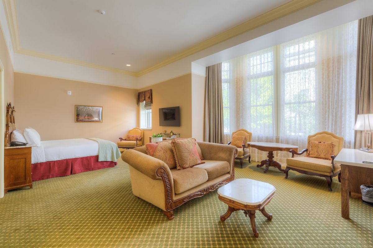 Spacious hotel room with a king bed, sofa, armchairs, and a large window overlooking the outdoors; neutral tones.