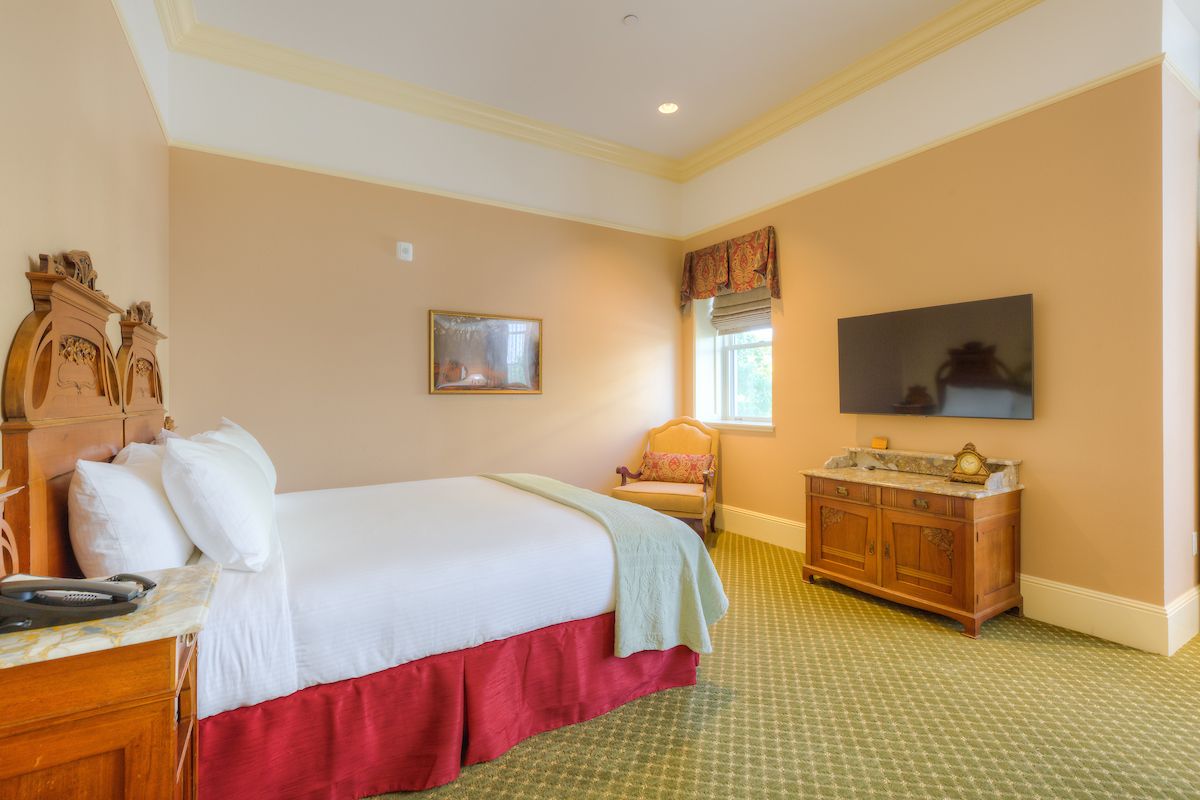 A hotel room with a bed, TV, and antique furniture. Beige walls, red bed skirt, and green carpet.