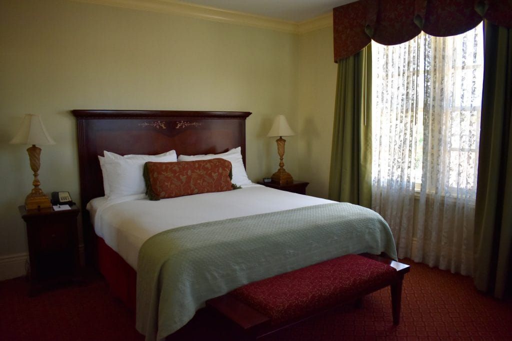 Hotel bedroom with a bed, nightstands, window, and a red bench. The bed has white linens, a green throw.