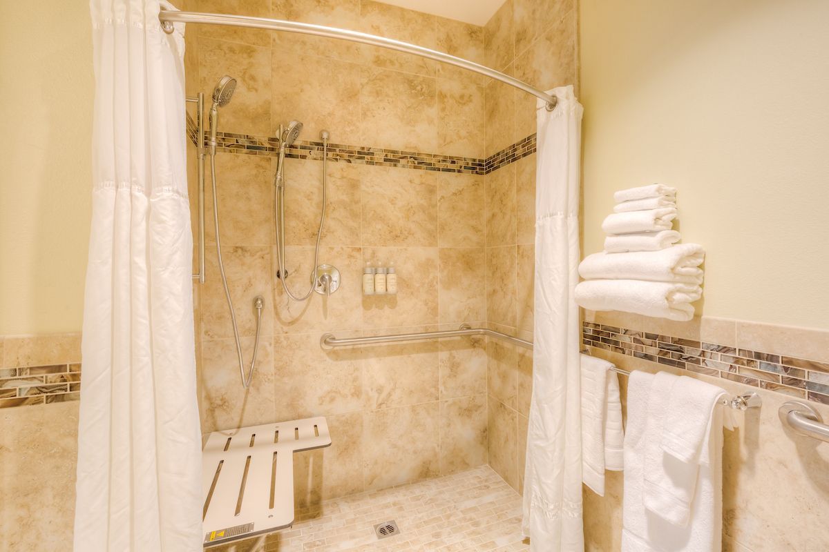Accessible hotel shower with beige tiles, a shower bench, grab bar, and stacked white towels.