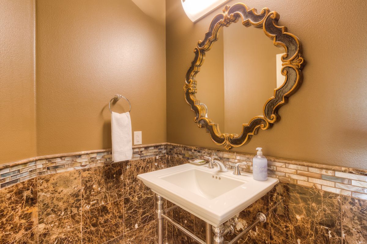 A luxurious powder room with a  sink, ornate gold-framed mirror, and marble walls. A towel hangs on a chrome towel rack.