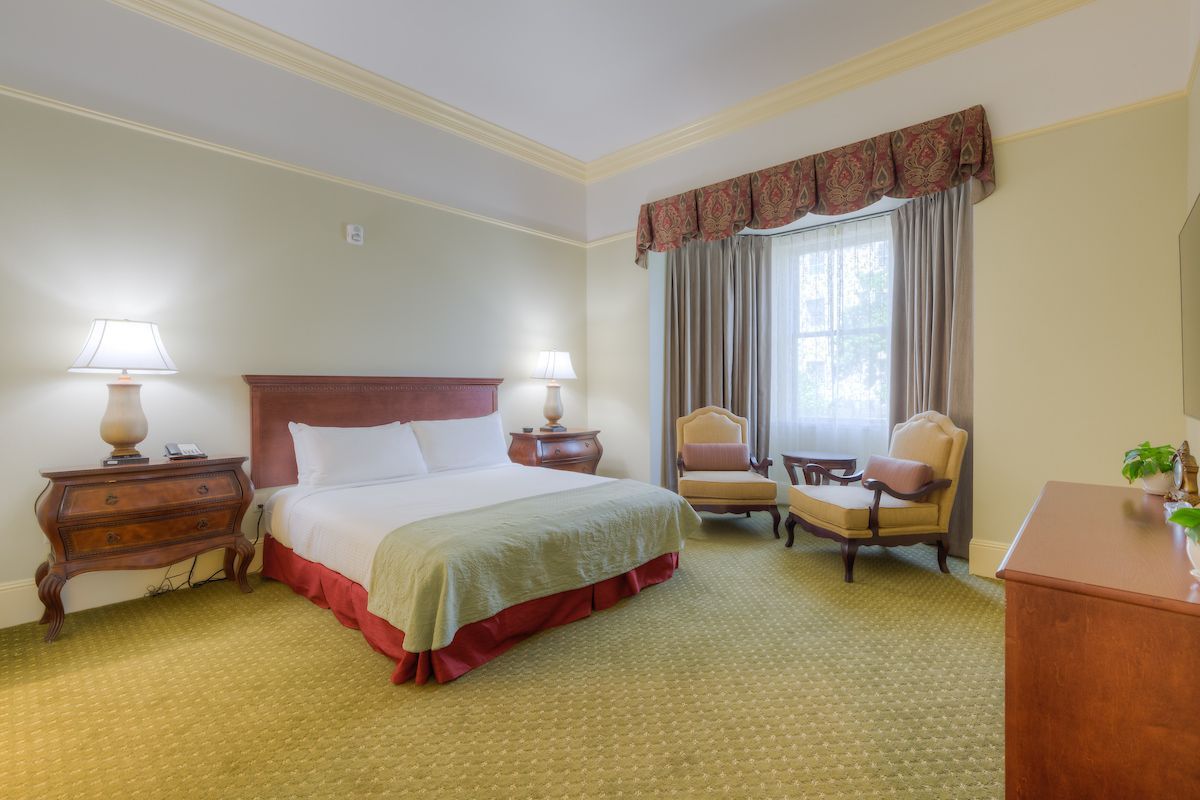 Hotel room with a bed, two chairs, a window with curtains, and two bedside tables with lamps. The room has a green carpet.