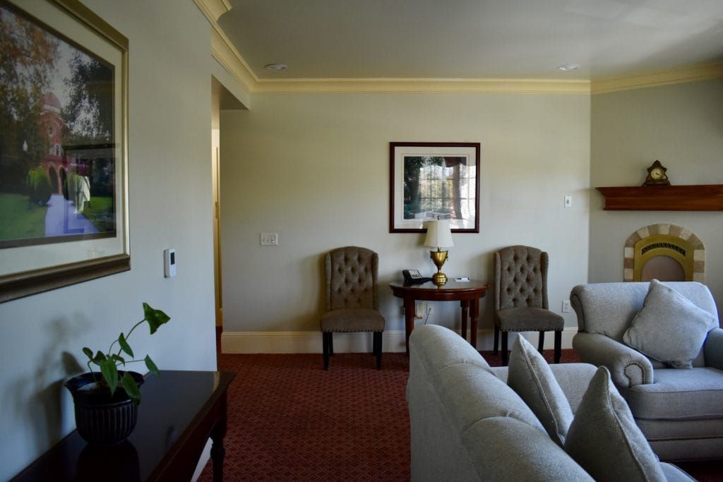 A living room with neutral walls, red patterned carpet, and a fireplace. There are two tufted chairs and a loveseat.
