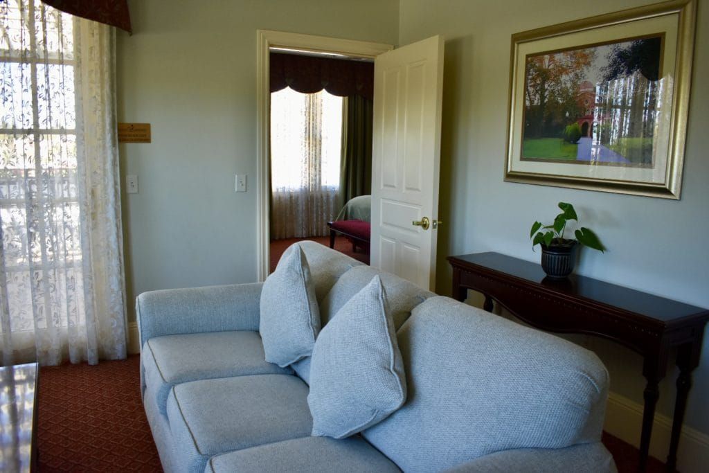A light gray couch sits in front of a window with lace curtains. A door leads to another room.