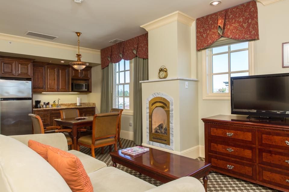Interior of a hotel suite with a kitchenette, dining area, living room, and fireplace. The decor is traditional.