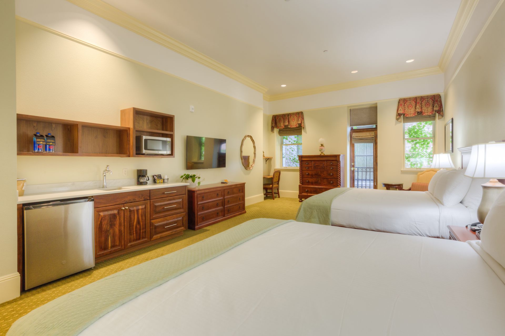 A hotel room with two queen beds, a kitchenette, and a balcony. Warm tones, hardwood furniture.