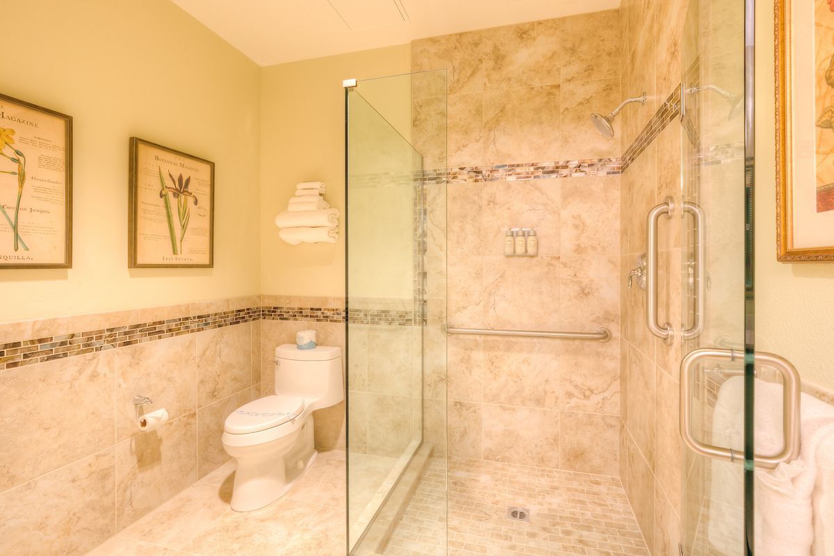 Bathroom with glass shower, toilet, and beige marble tile. Two floral prints hang on a light yellow wall.