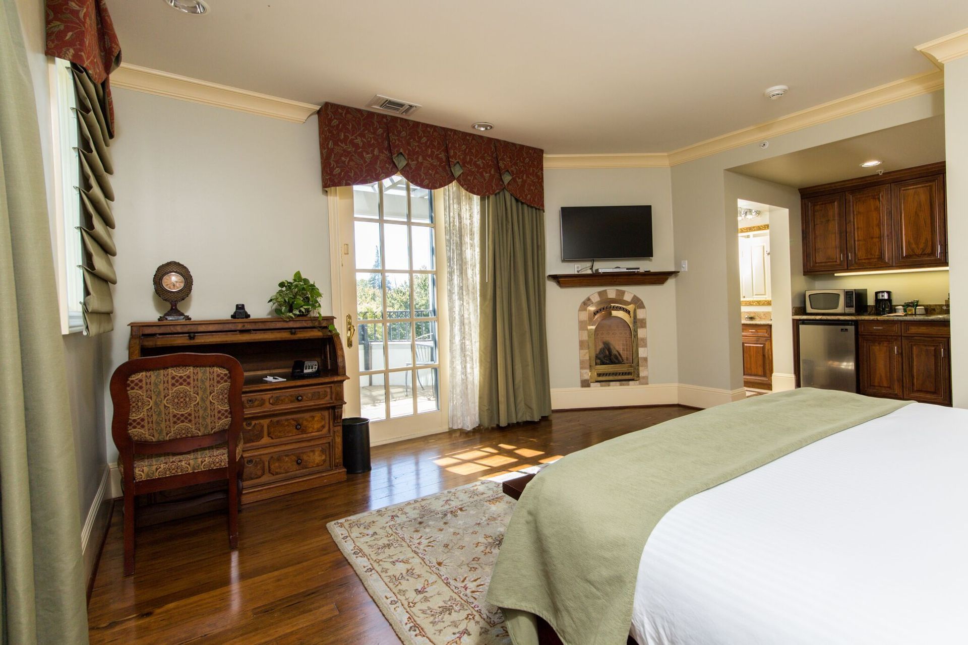 Bedroom with a bed, fireplace, desk, and a small kitchenette. A doorway leads to a balcony, with light streaming in.