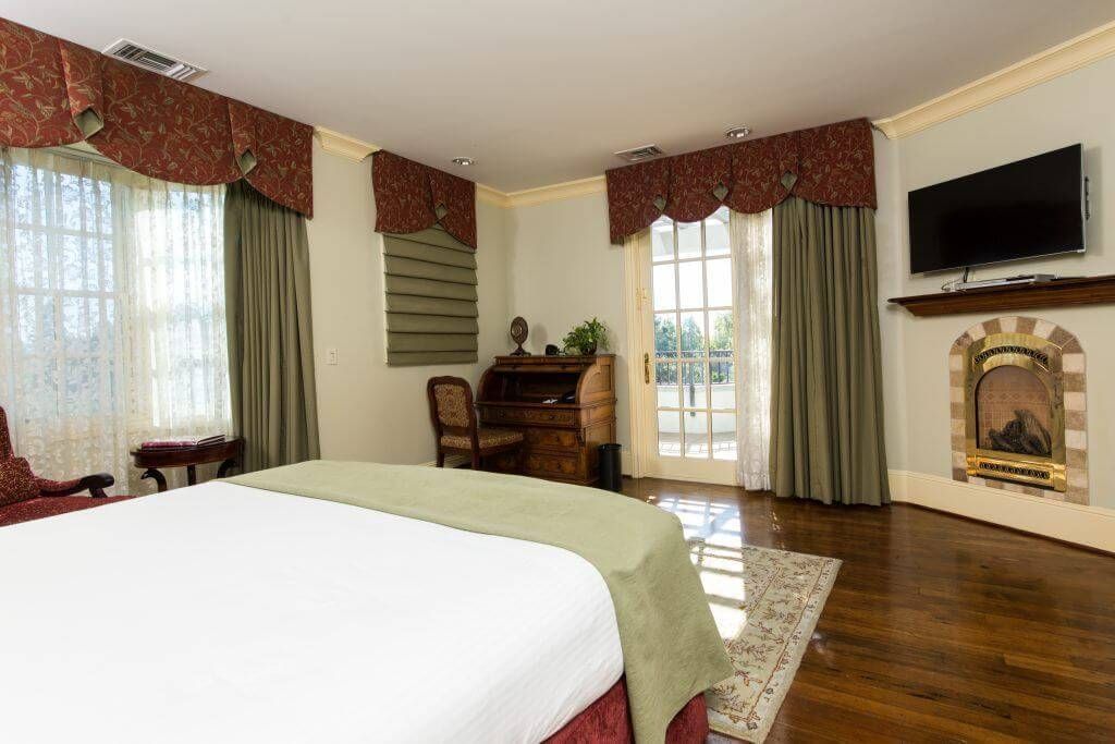 Spacious bedroom with a king-size bed, fireplace, and large windows. Red curtains, light green bed cover, and antique desk.