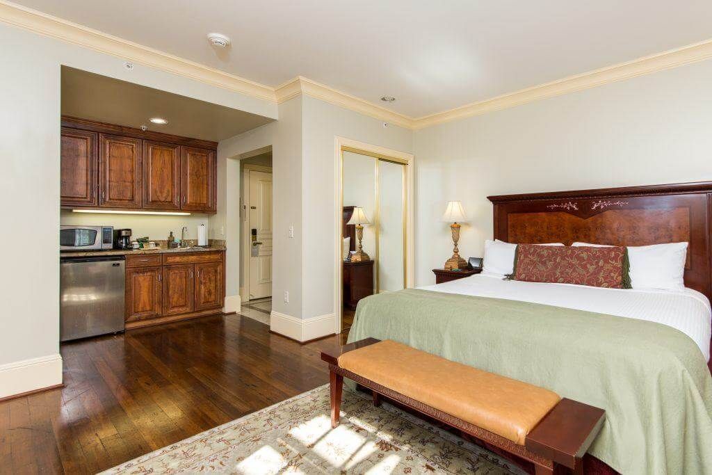 A hotel room with a bed, mini-kitchen, and a door to a bathroom. Wooden floors, cabinets, and a green bedspread.