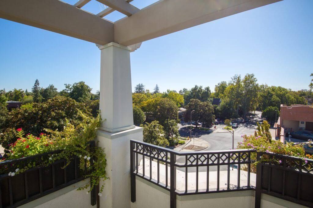 Balcony overlooking a tree-lined street, with a white column, trellis, and dark brown railing under a bright blue sky.