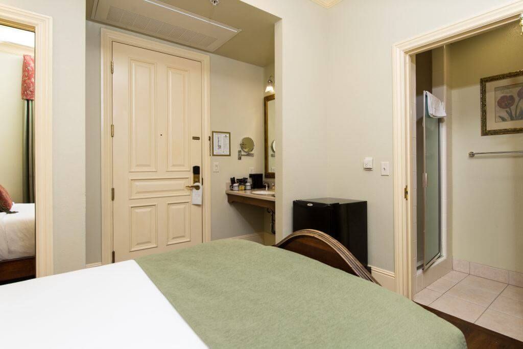 A hotel room with a bed, bathroom, and doorway to another room. The bed has a green cover, and the walls are light green.