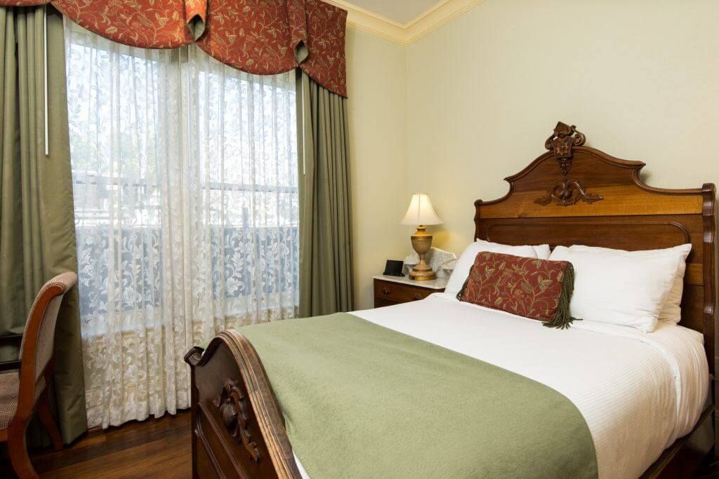 A hotel bedroom with a made bed, window with sheer curtains, desk, and decorative wooden headboard. Green and red accents.