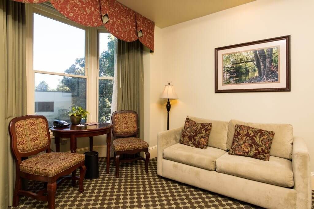 A hotel room with a beige sofa, two patterned chairs, a small table, and a window with green curtains.