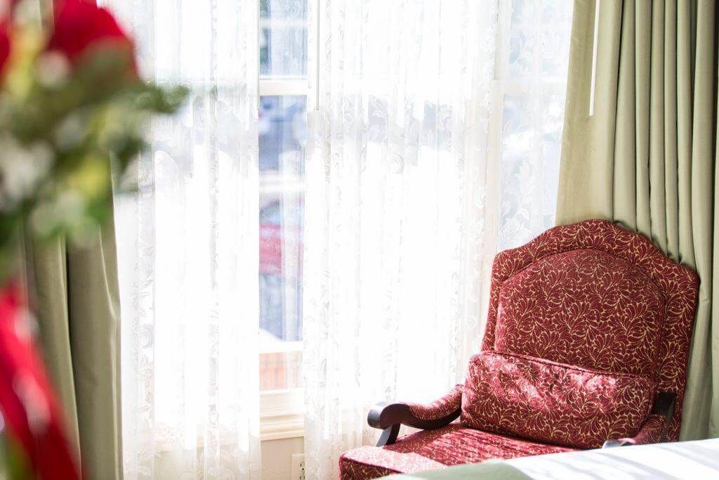 A maroon armchair with a patterned seat sits near a window with sheer curtains.