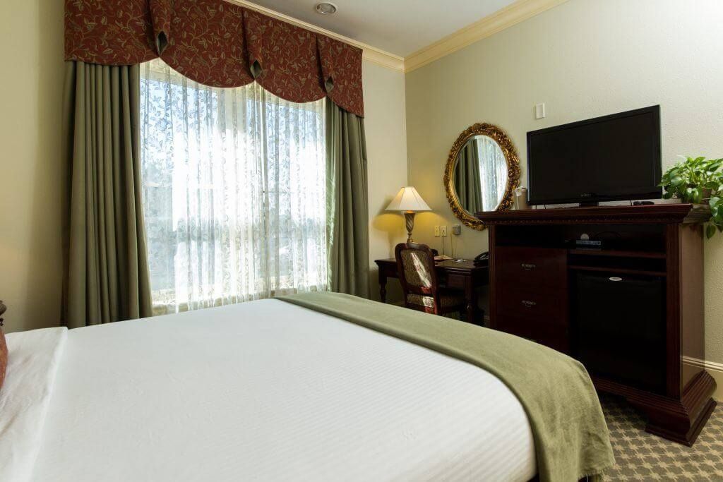 Hotel room with a bed, television, desk, and window with curtains. Room is decorated in earth tones.