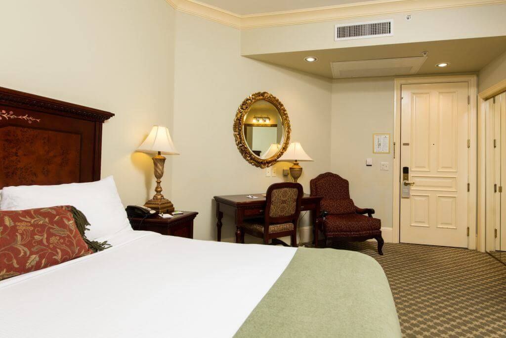 Hotel room with a bed, desk, and armchair. The room has beige walls and patterned carpet.