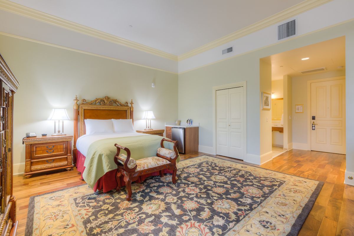 Hotel bedroom with wooden furniture, a large patterned rug, and a view into a bathroom.