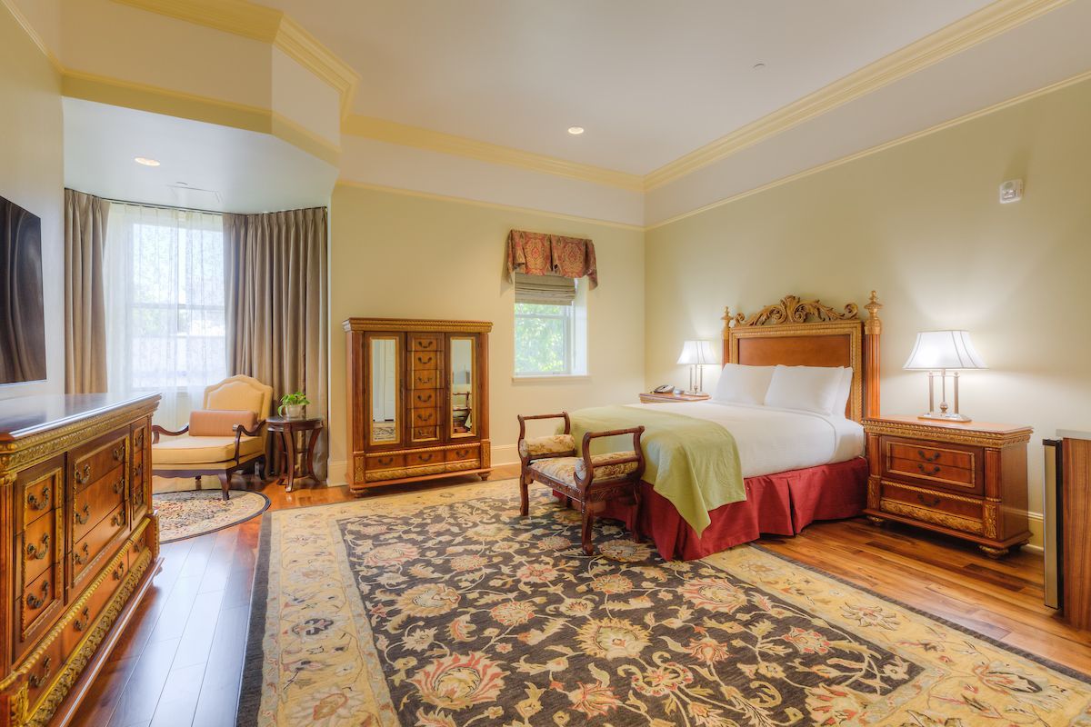 A hotel room with a bed, dresser, and sitting area. The room has a large area rug and wood furniture.