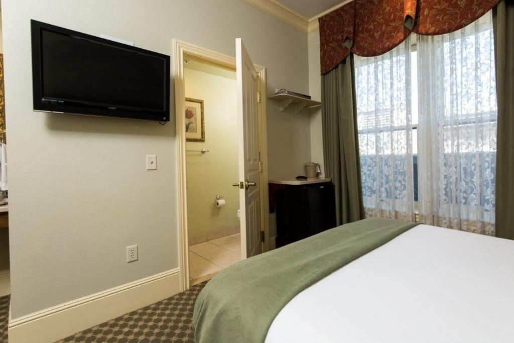 Hotel room interior with a bed, TV, bathroom, and window. Green and brown accents throughout.