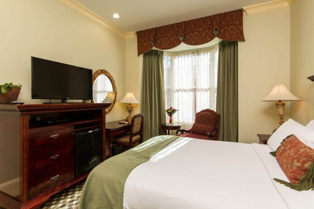 A hotel room with a bed, TV, desk, and bay window with curtains. The room is decorated in shades of gold, green, and brown.