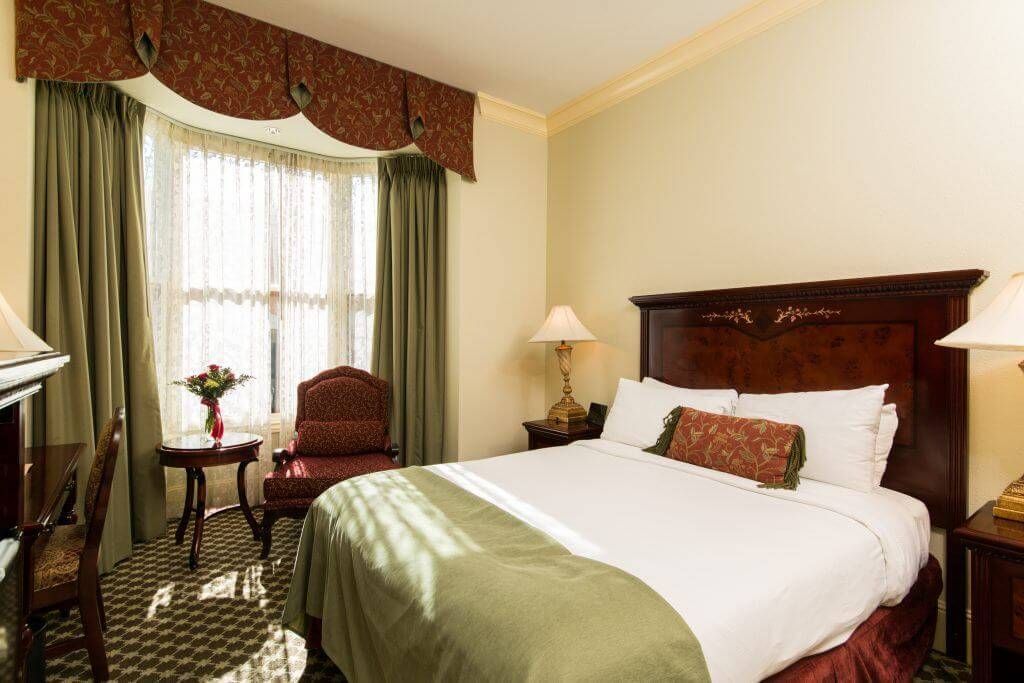 Hotel bedroom with a queen-sized bed, a seating area near a bay window, and warm lighting.