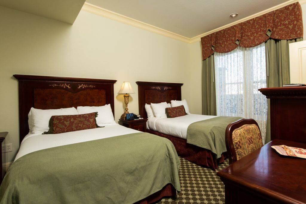 Hotel room with two beds, brown headboards, green bedspreads, and a patterned carpet. A window is visible in the background.