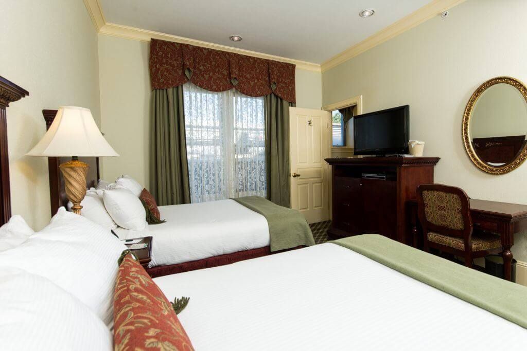 A hotel room with two beds, a desk, and a TV. Cream walls, a decorative window treatment, and warm-toned bedding.