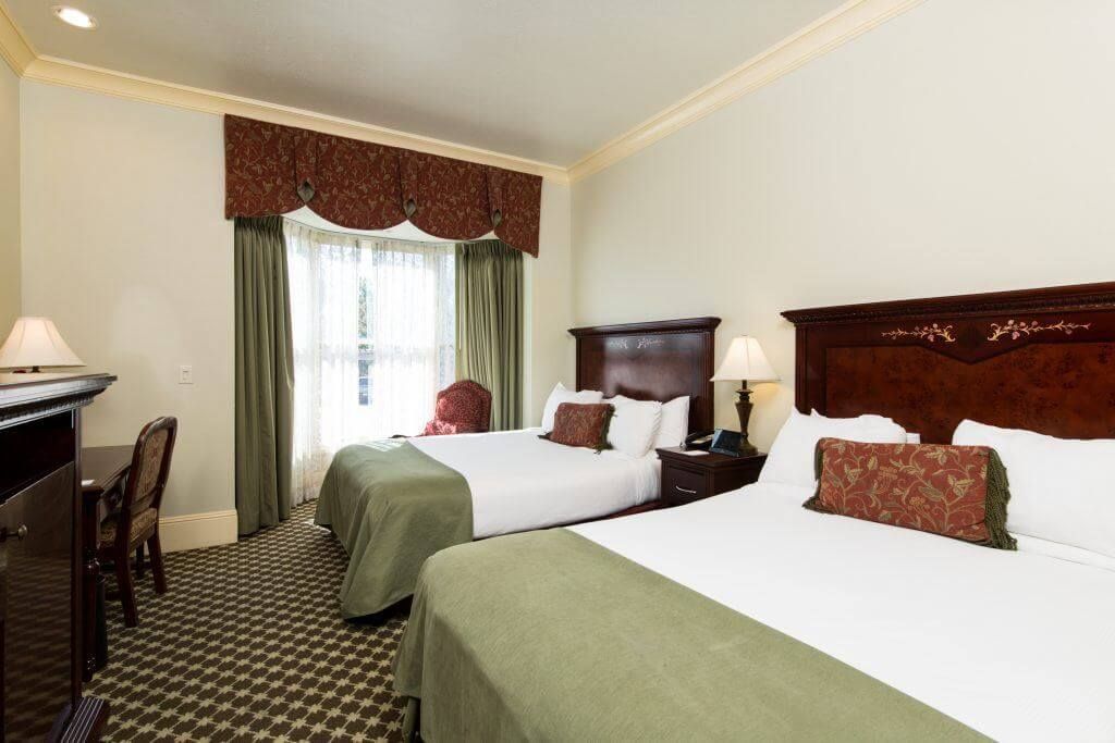 A hotel room with two double beds, a desk, and a window with green curtains. 