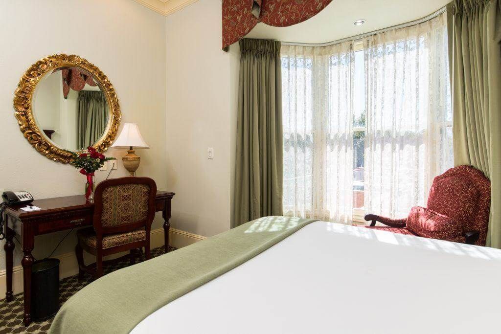 Elegant hotel room with a large bed, desk with mirror, and window with a cozy armchair. 