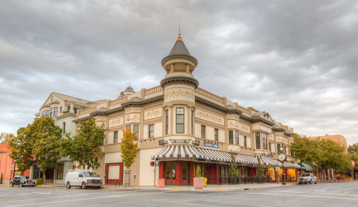 Corner view of a historic, ornate building with a turret, awnings, and storefronts; cloudy sky overhead.