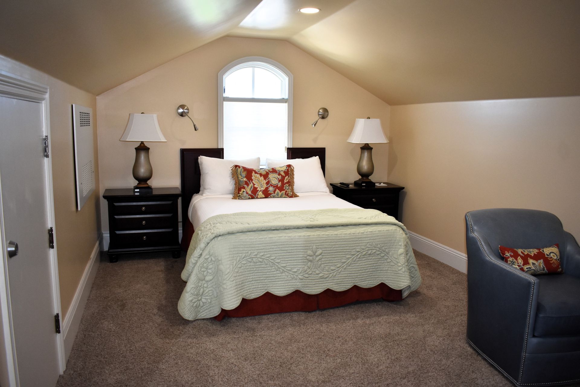 Bedroom with bed, nightstands, armchair, and arched window in a sloped-ceiling room. Warm beige walls and a beige carpet.