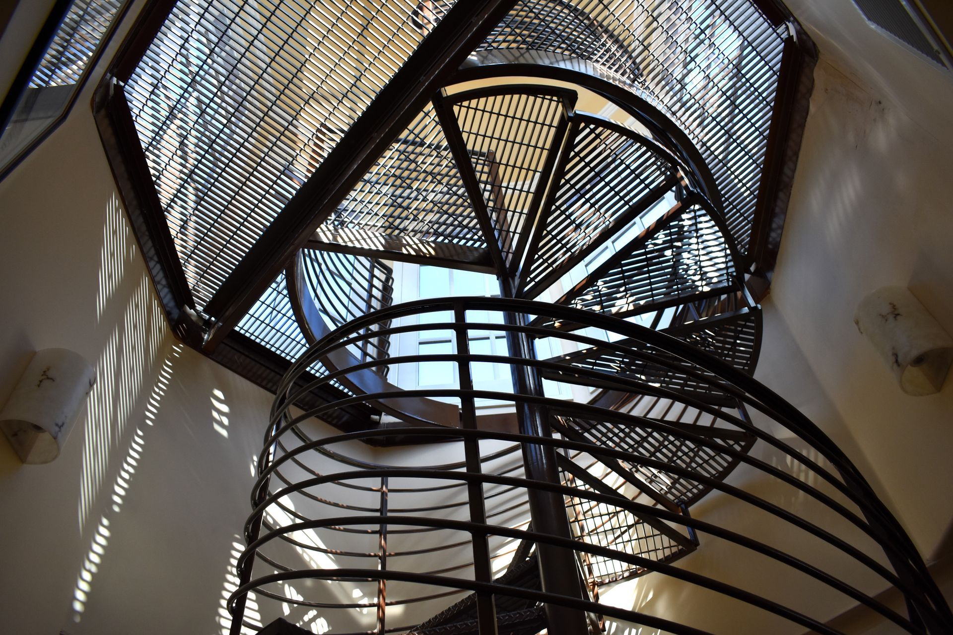 Spiral metal staircase leading up to a circular opening with a glass grid ceiling, viewed from below. 