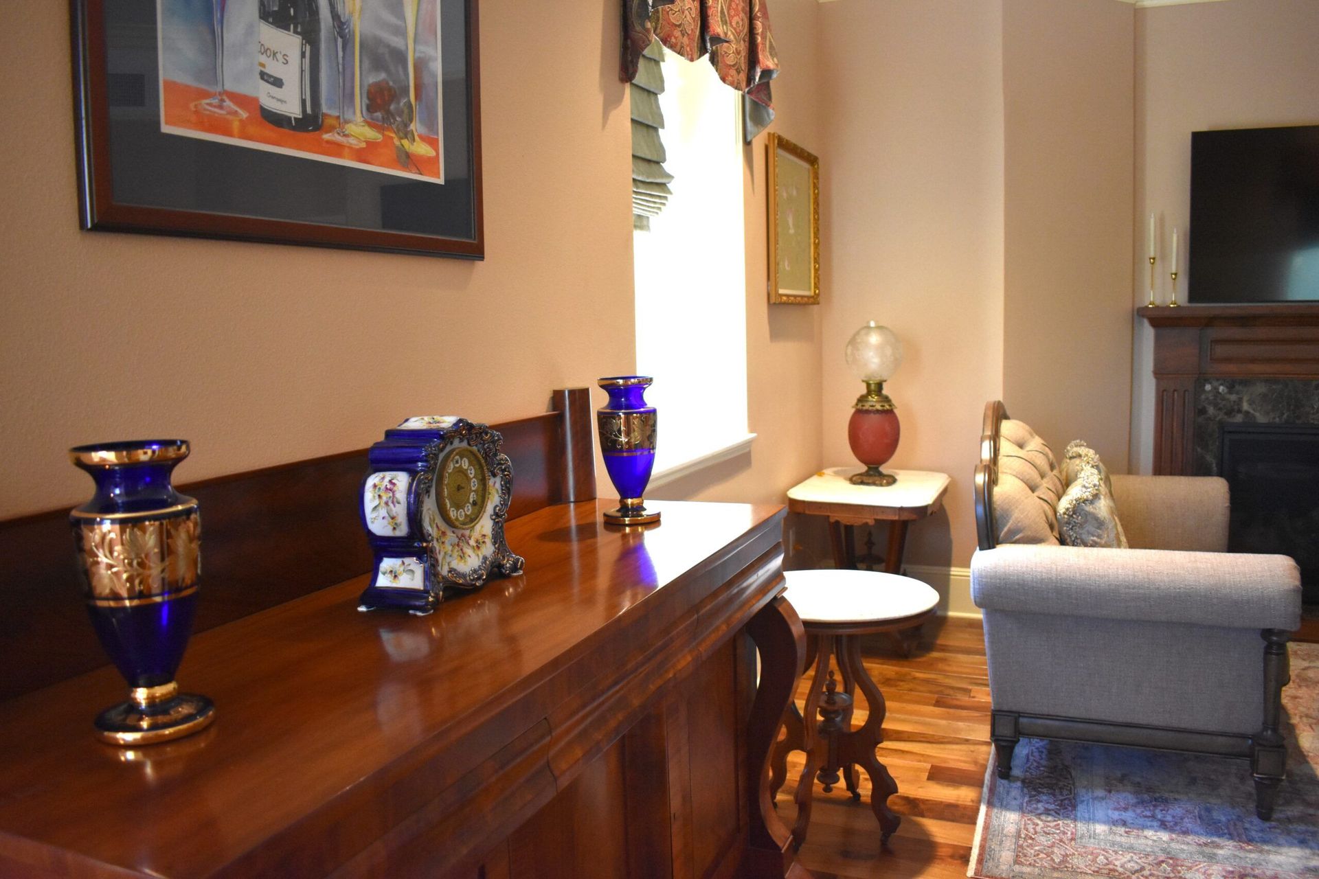 A room with a long wooden sideboard featuring blue and gold vases and a clock. A window, small table, and sofa are visible.