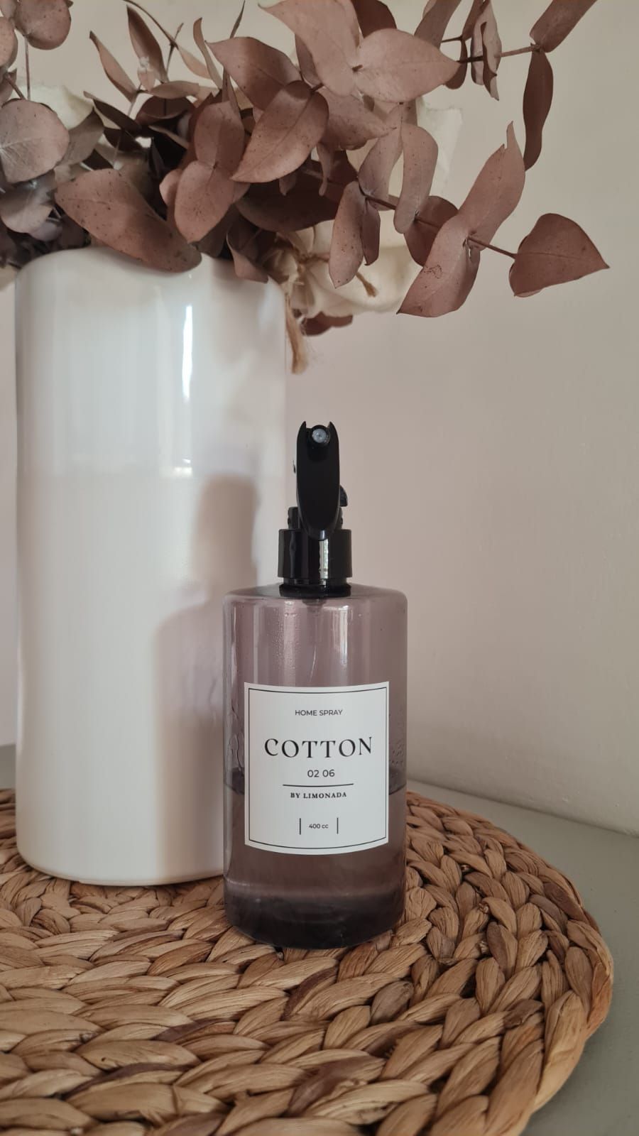 Una botella con atomizador para ambientes y un jarrón blanco con hojas marrones sobre un mantel tejido.