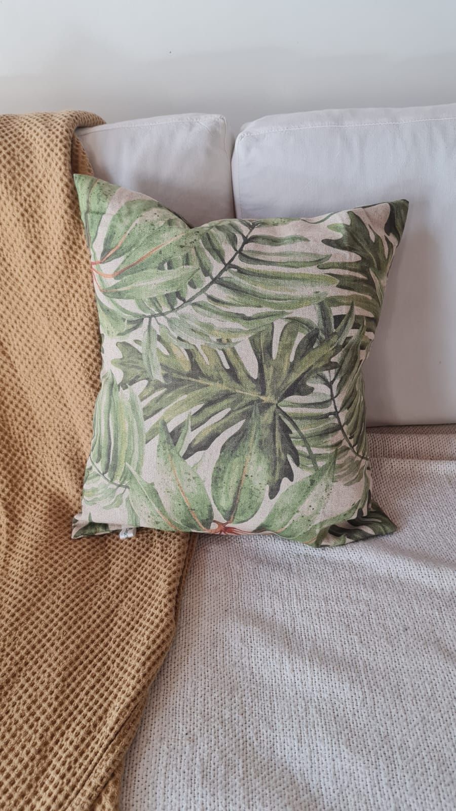 Cojín con estampado de hojas tropicales verdes sobre un sofá blanco, con una manta color tostado y una manta de punto blanca.