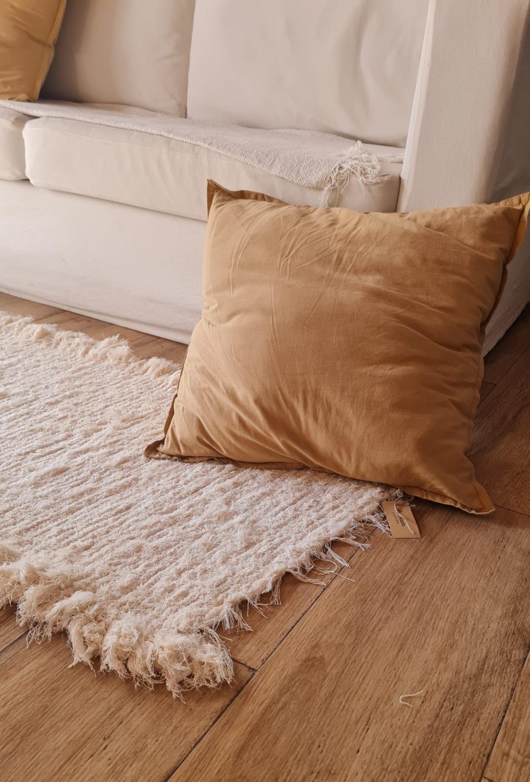 Sofá blanco, almohada color canela y alfombra con flecos blancos sobre un piso de madera.