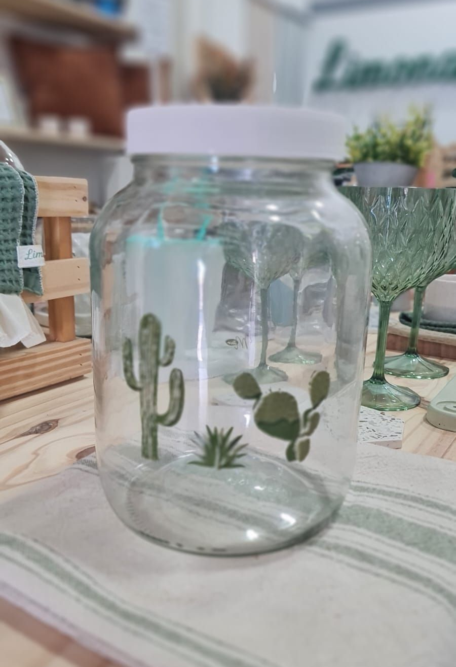 Tarro de cristal con tapa dorada y hojas de monstera pintadas de verde sobre superficie de madera.