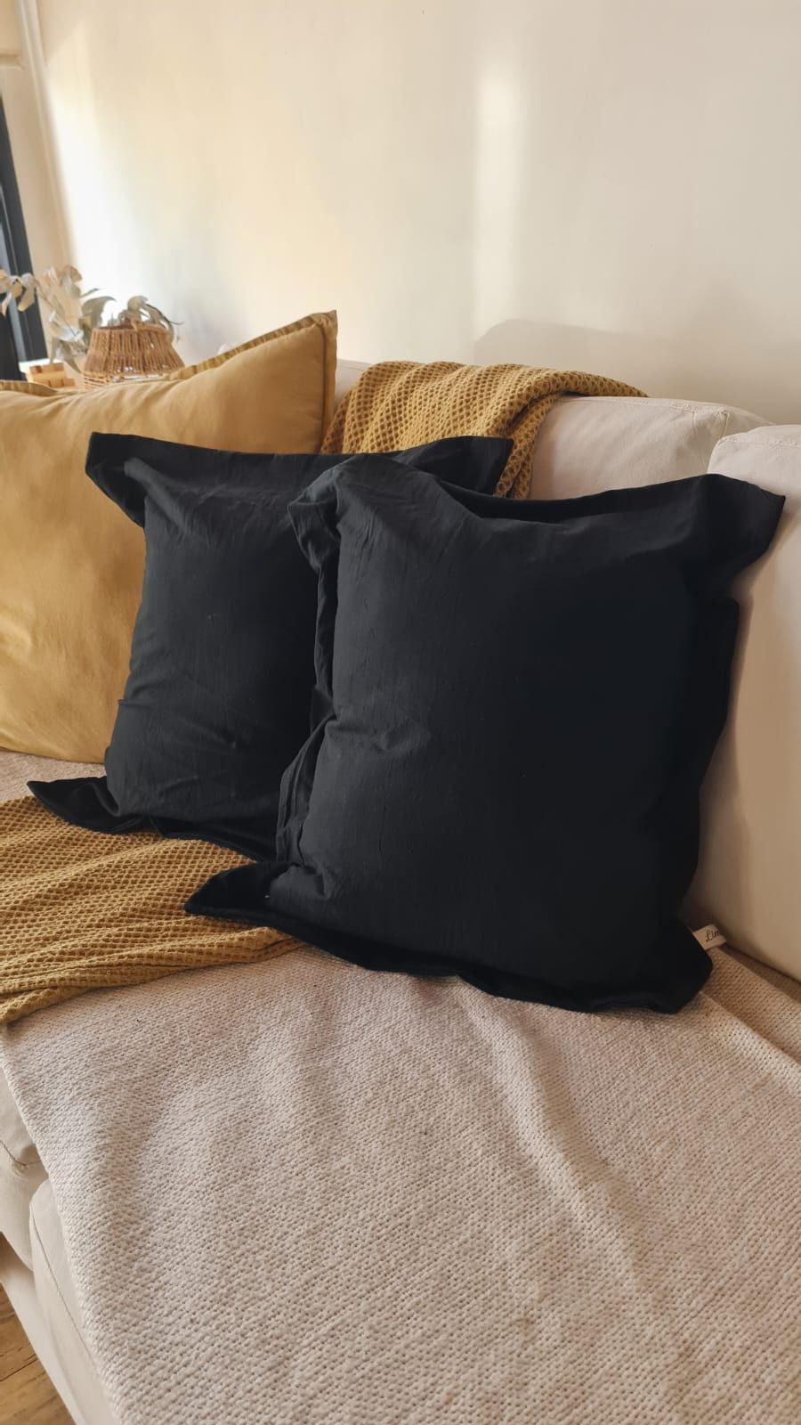 Cojines negros y dorados sobre un sofá blanco con una manta beige texturizada. Ambiente con luz natural.