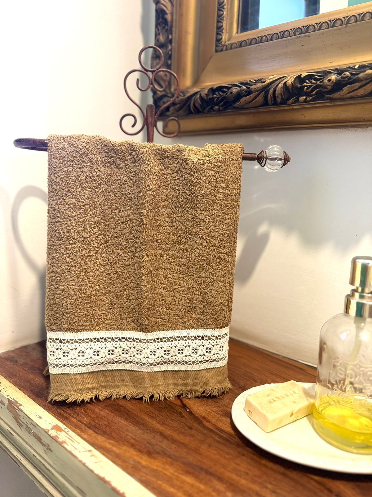 Toalla de arpillera marrón con ribete de encaje blanco sobre un toallero decorativo de cobre en un baño.