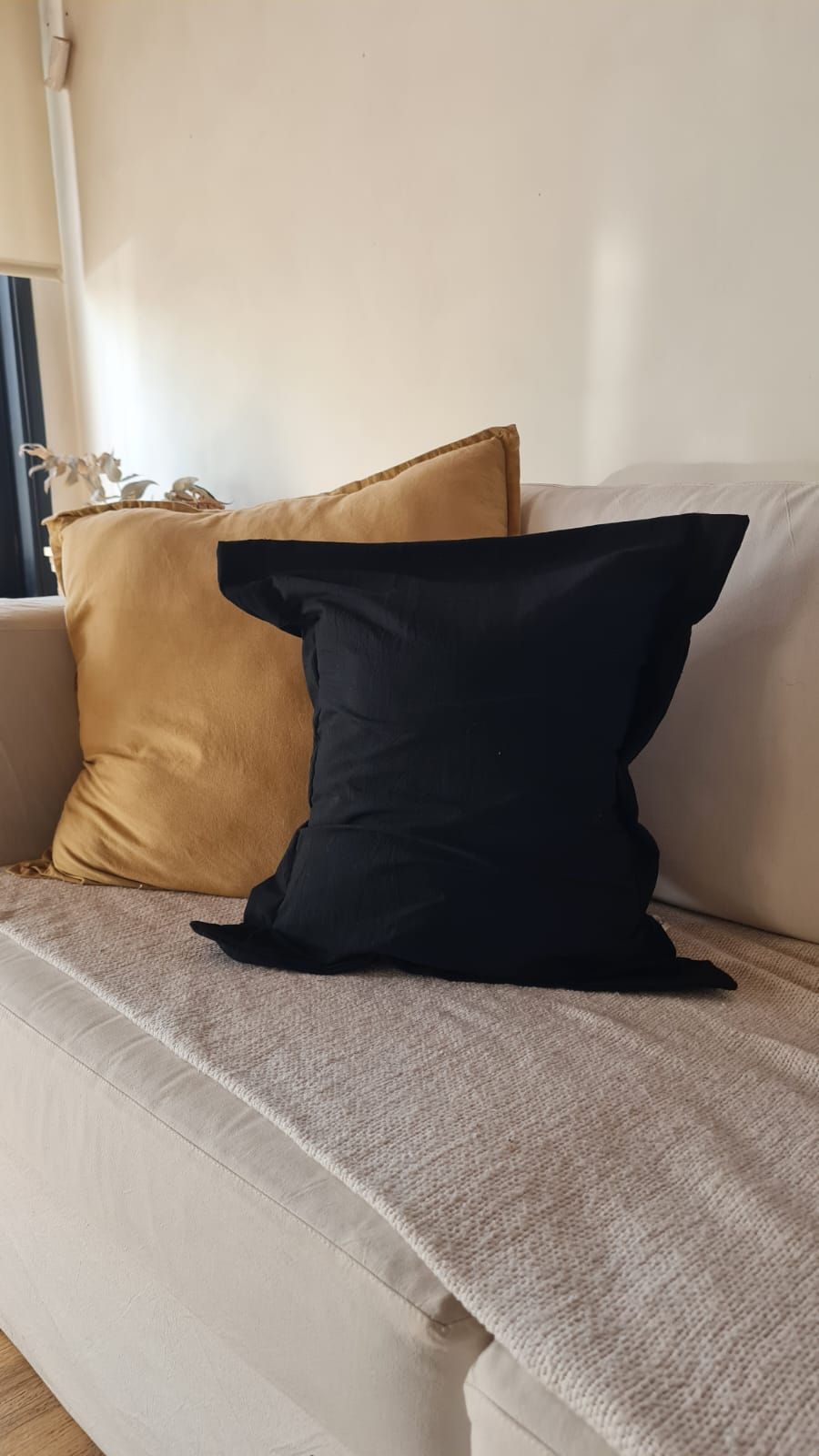 Cojines negros y dorados sobre un sofá blanco con una manta beige texturizada. Ambiente con luz natural.